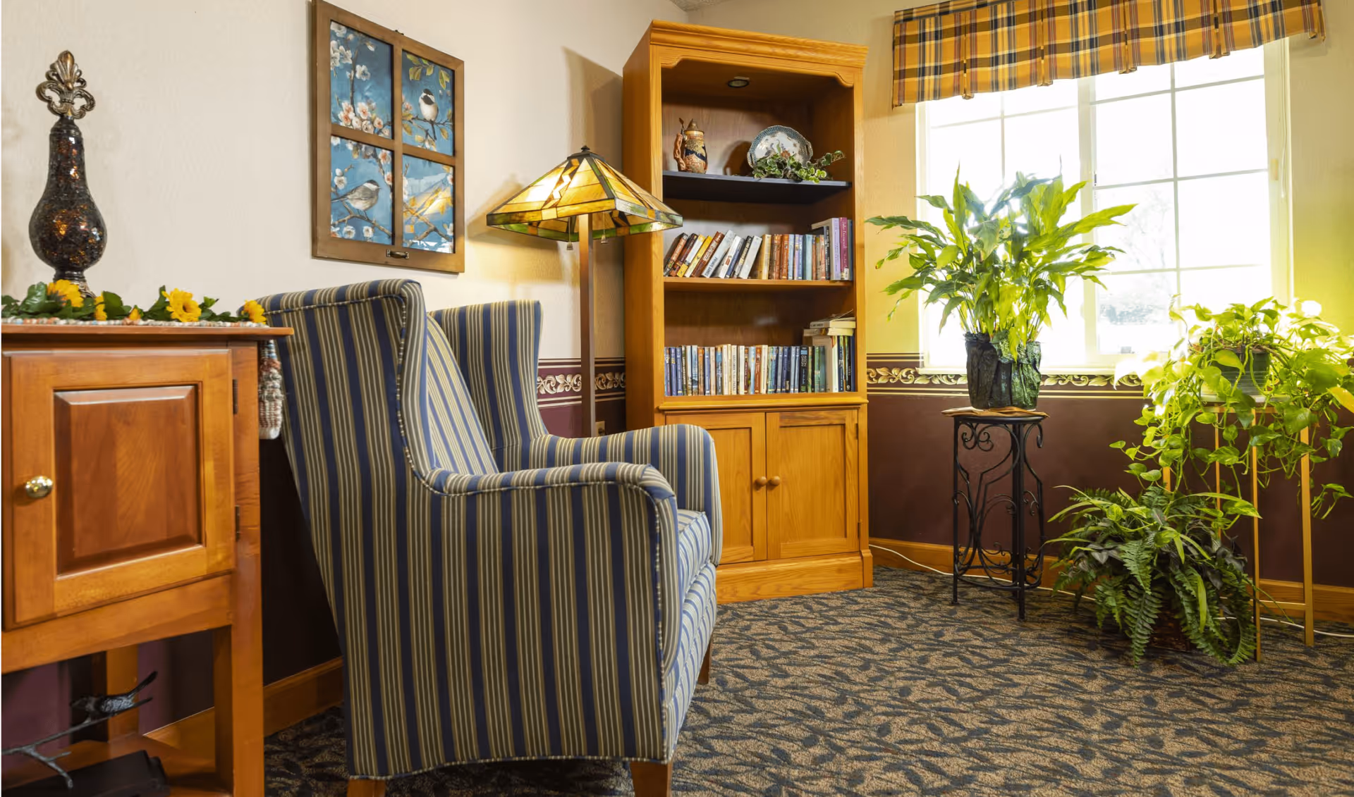 A cozy corner of a living room with a striped armchair, a wooden bookshelf filled with books and decorative items, a Tiffany-style floor lamp, a wooden side table with a decorative vase and sunflowers, and several green potted plants near a window with plaid valance curtains.
