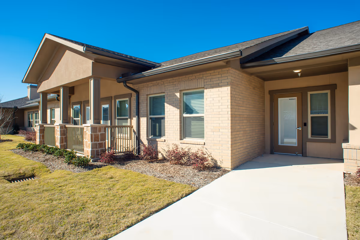 Single-story beige brick senior living building with a covered front porch and manicured lawn