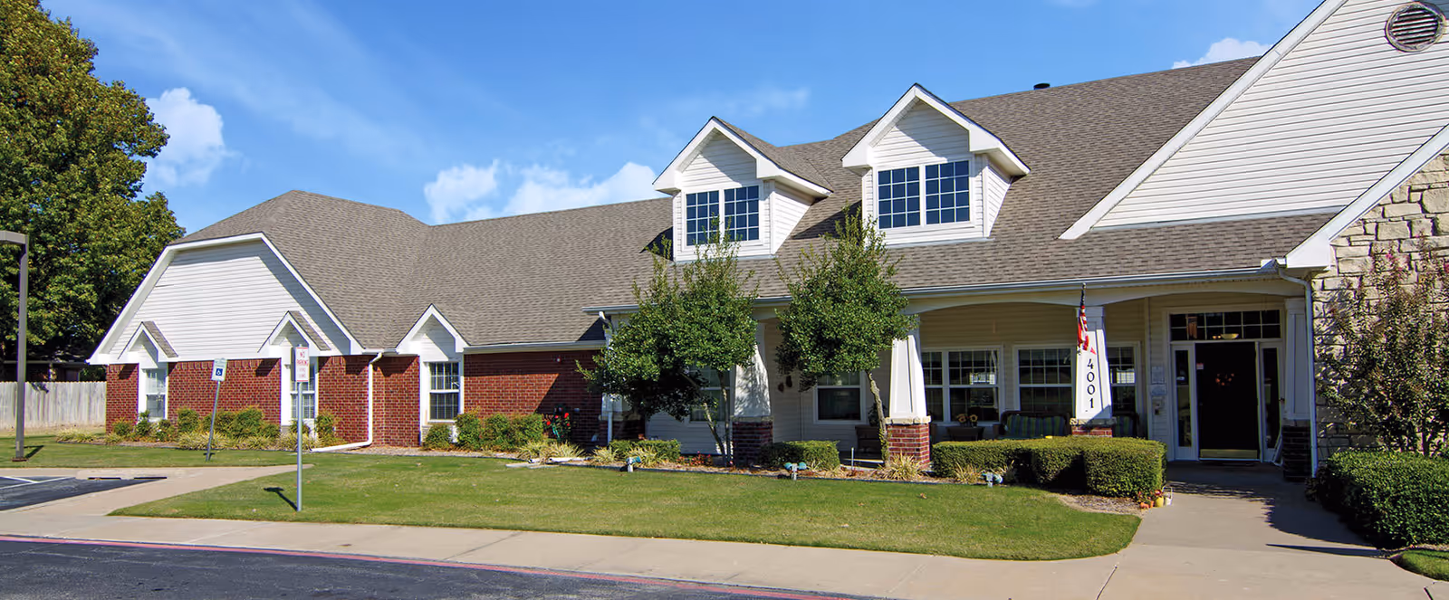 Exterior view of a single-story senior living facility building with a combination of brick and white siding, multiple windows, a covered entrance with columns, and a well-maintained lawn and shrubs under a blue sky with some clouds.