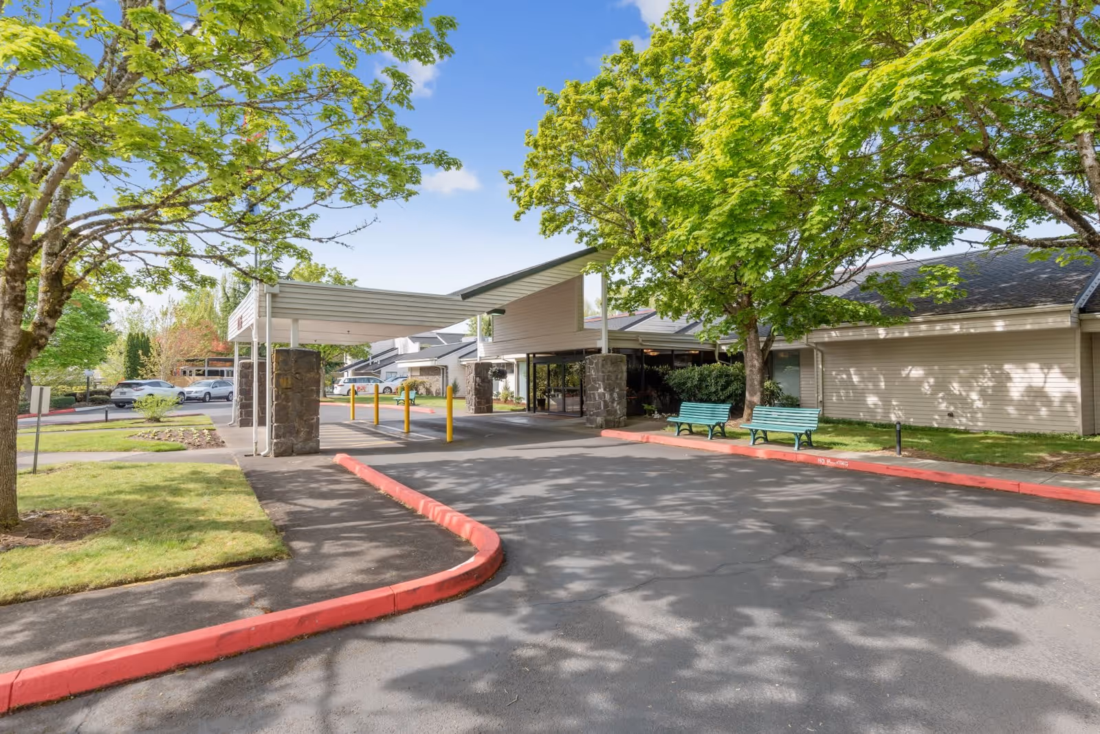 Entrance driveway to a retirement community building with a covered drop-off area supported by stone pillars, surrounded by green trees and grass, with two green benches on the right side and several parked cars in the background under a clear blue sky.