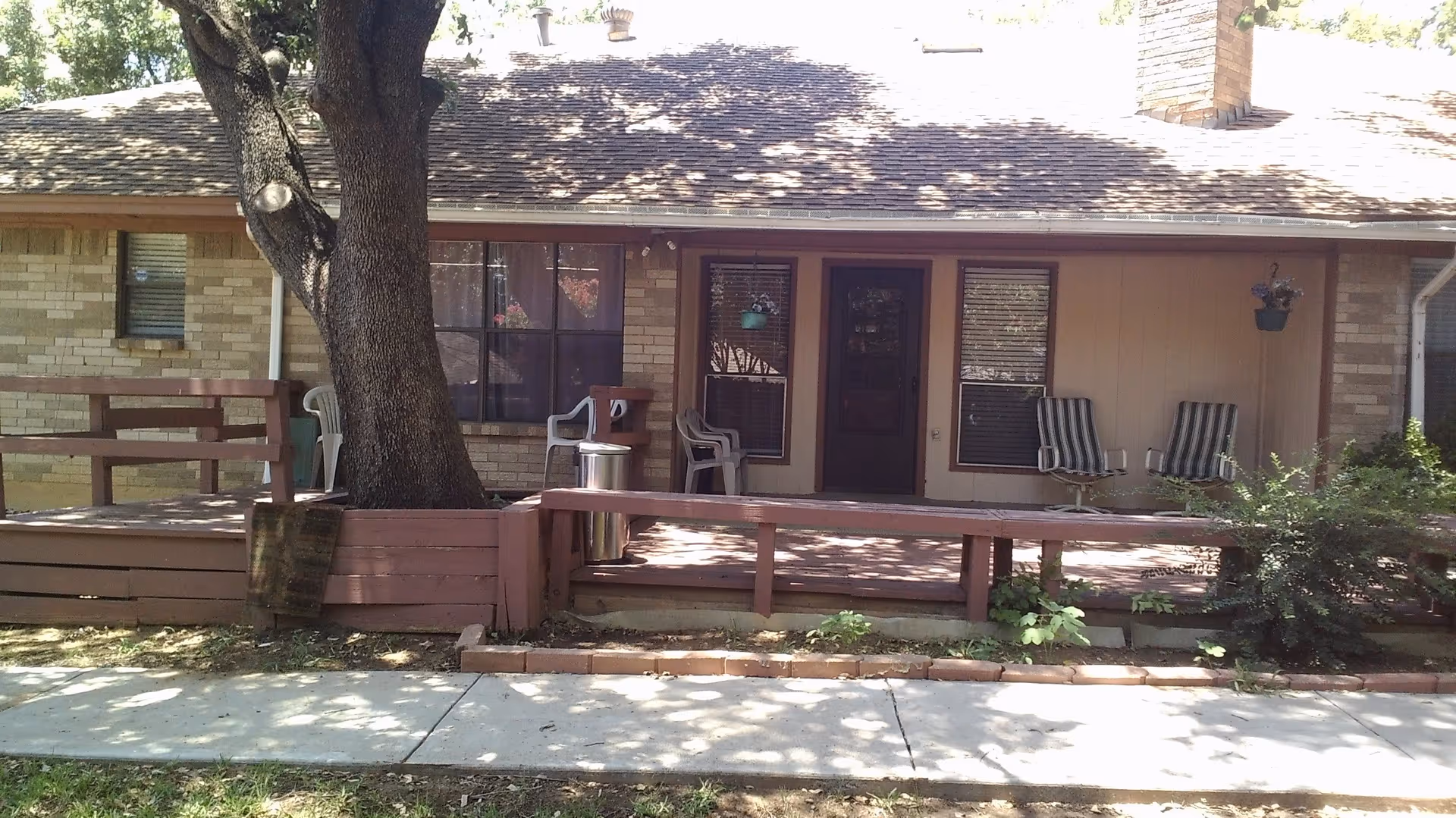 Front exterior of a single-story building with a wooden porch, chairs, hanging plants, and a large tree in front.