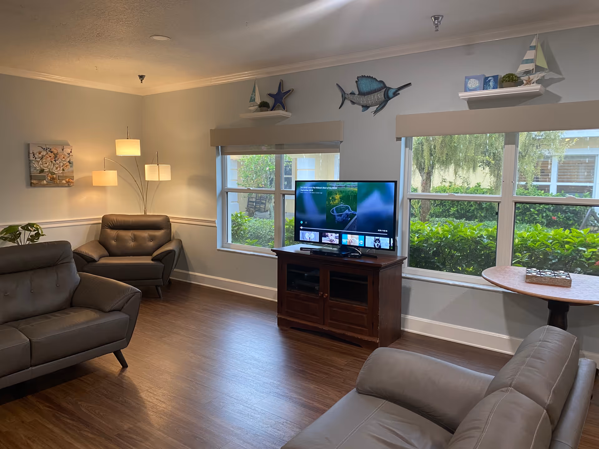 A cozy living room in Cypress Creek Assisted Living featuring three gray leather armchairs, a wooden TV stand with a flat-screen TV, large windows with views of greenery outside, a round wooden side table, a floor lamp with three lights, and decorative wall shelves with nautical-themed items.