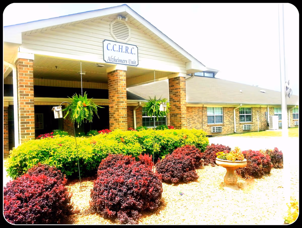 Exterior view of the Cherokee County Health and Rehabilitation Center Alzheimer's Unit building entrance with brick pillars, a covered porch, and well-maintained landscaping featuring green and red bushes, hanging plants, and a decorative birdbath.