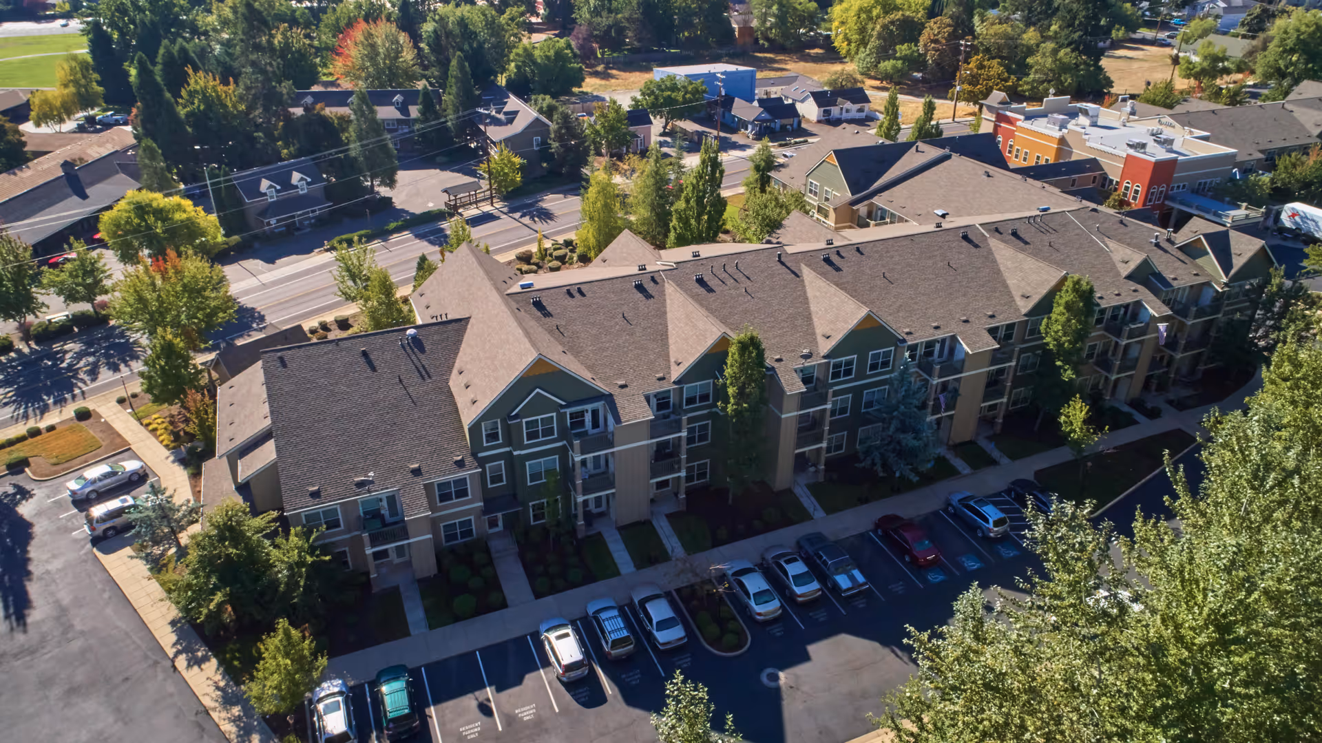 Aerial view of a multi-story residential building complex surrounded by trees and parking spaces with several cars parked. The buildings have pitched roofs and multiple windows, with a road and more greenery visible in the background.