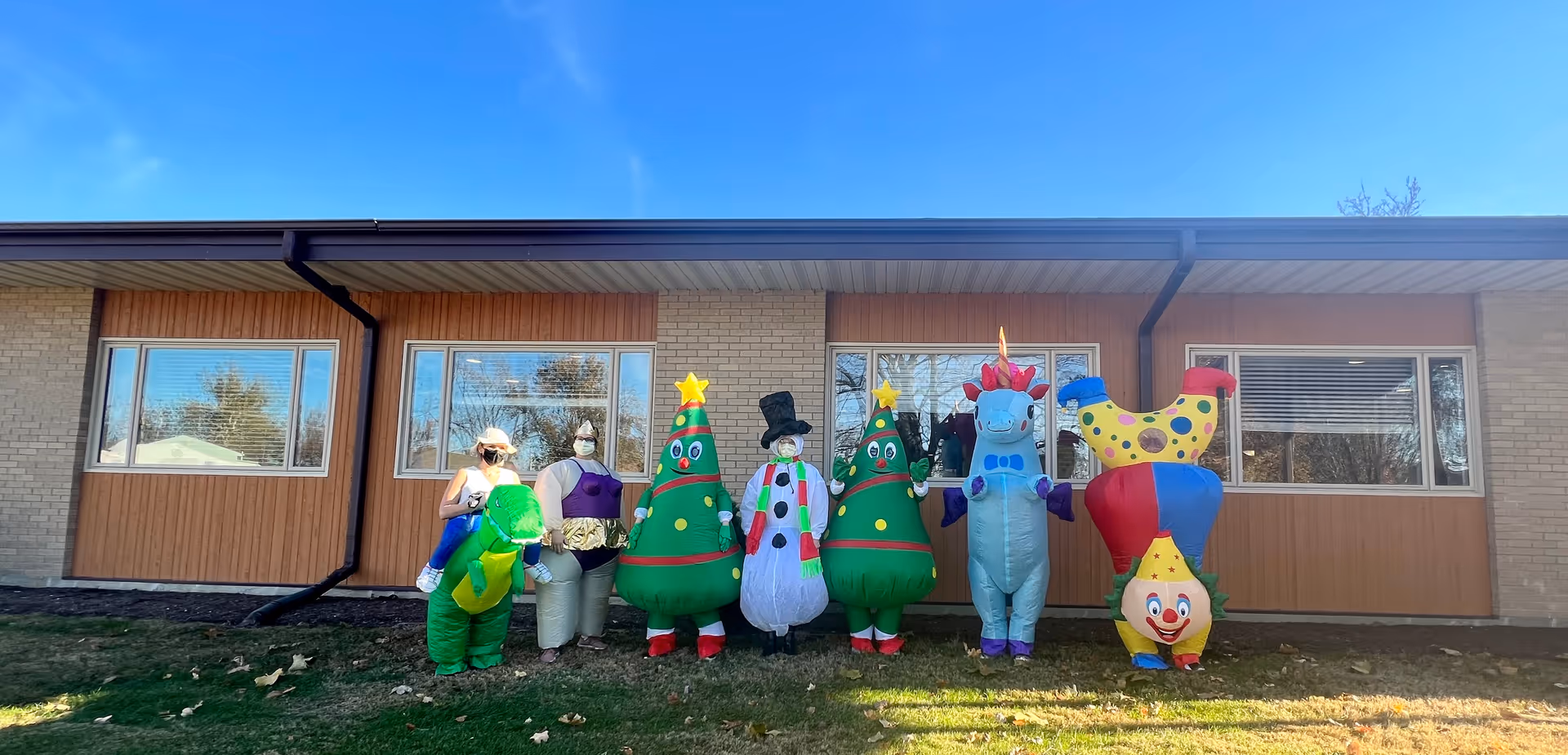 Seven people dressed in colorful inflatable costumes including a dinosaur, sumo wrestler, two Christmas trees, a snowman, a unicorn, and a clown standing outside in front of a single-story building with large windows under a clear blue sky.