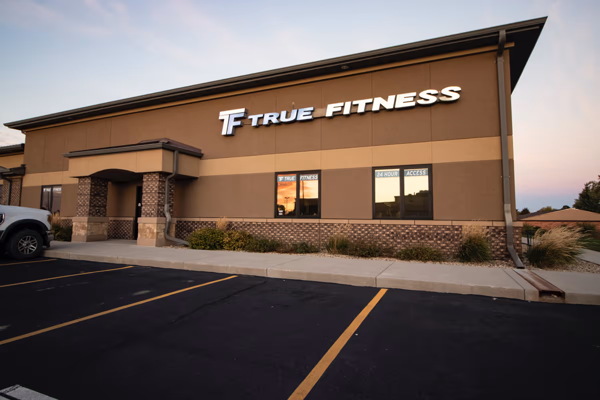 Exterior of a tan building with a True Fitness sign and a parking lot.