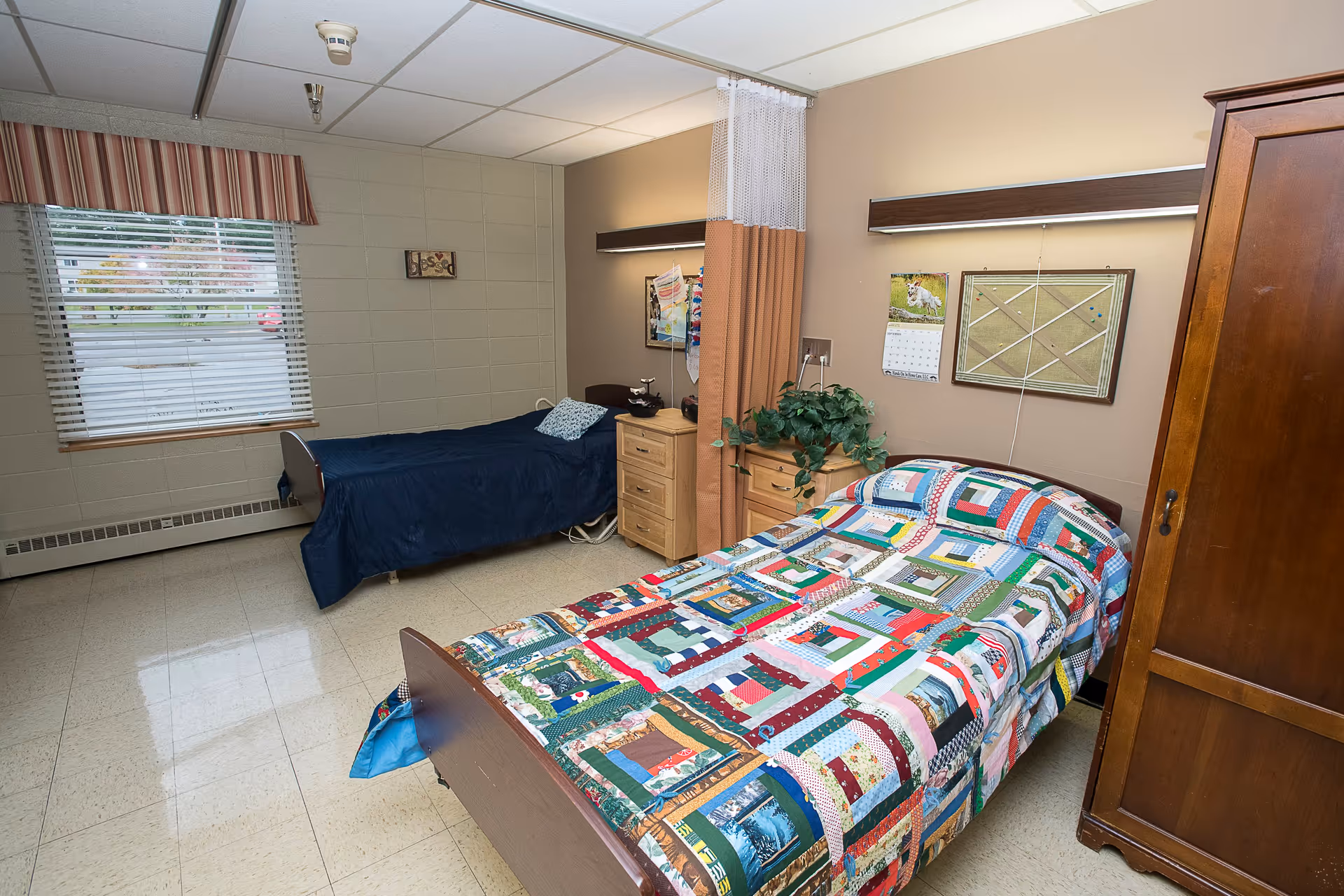 A shared bedroom in a senior living facility with two single beds. One bed has a colorful patchwork quilt, and the other has a dark blue bedspread. There is a wooden wardrobe, two small wooden dressers, a window with blinds and a striped valance, a calendar, a bulletin board, and a privacy curtain between the beds.