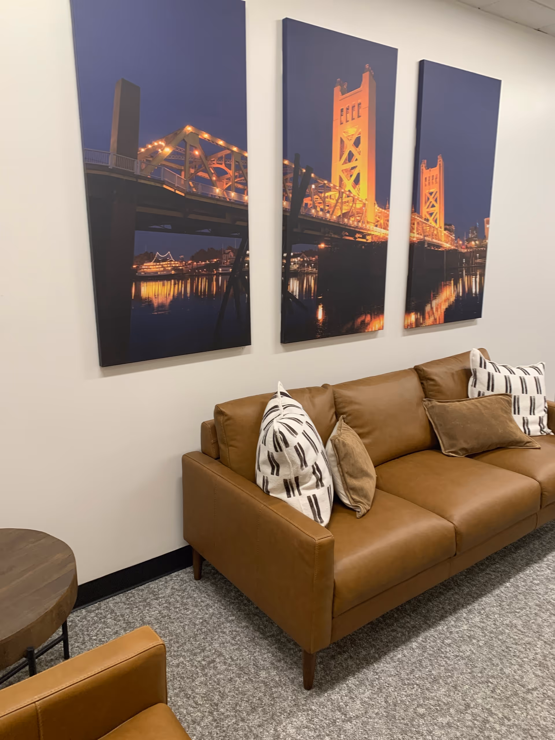 Brown leather sofa with patterned cushions beside three-panel bridge artwork in a living room.