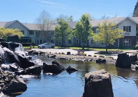 View of a senior living facility building named Jennings McCall Center with a pond and waterfall feature in the foreground, surrounded by rocks and trees under a clear blue sky.