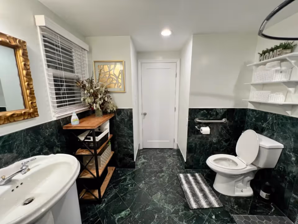 Bathroom with green marble floor and wainscoting, a white pedestal sink, open shelving, and a toilet with a grab bar.