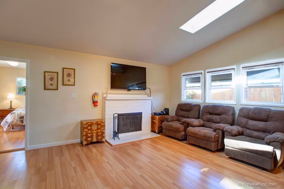 Bright living room with brown recliners, fireplace, mounted TV, windows, and a doorway to a bedroom.