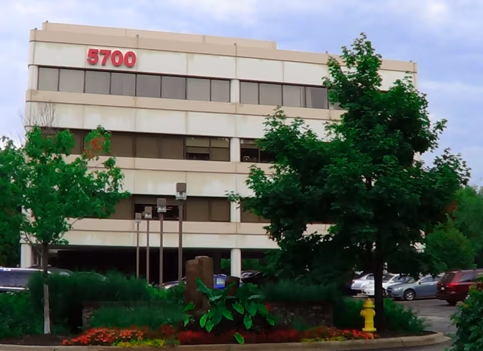 Beige, three-story building with large windows, greenery, and a front parking lot.