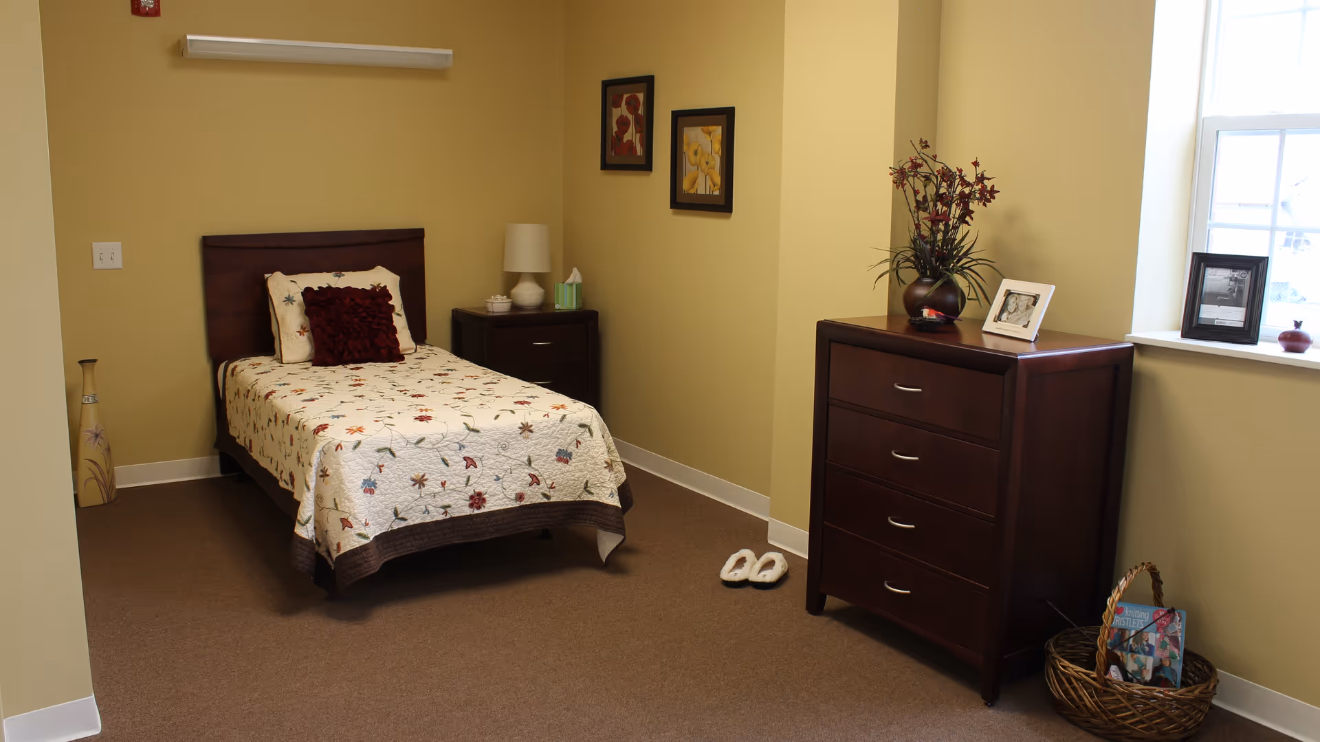 A cozy bedroom with a single bed covered in a floral quilt, a dark wooden headboard, and a burgundy pillow. Next to the bed is a nightstand with a lamp, tissue box, and small decorative items. On the right side of the room is a dark wooden dresser with a vase of flowers, framed photos, and a basket with magazines on the floor. The walls are painted light yellow, and there is a window letting in natural light.