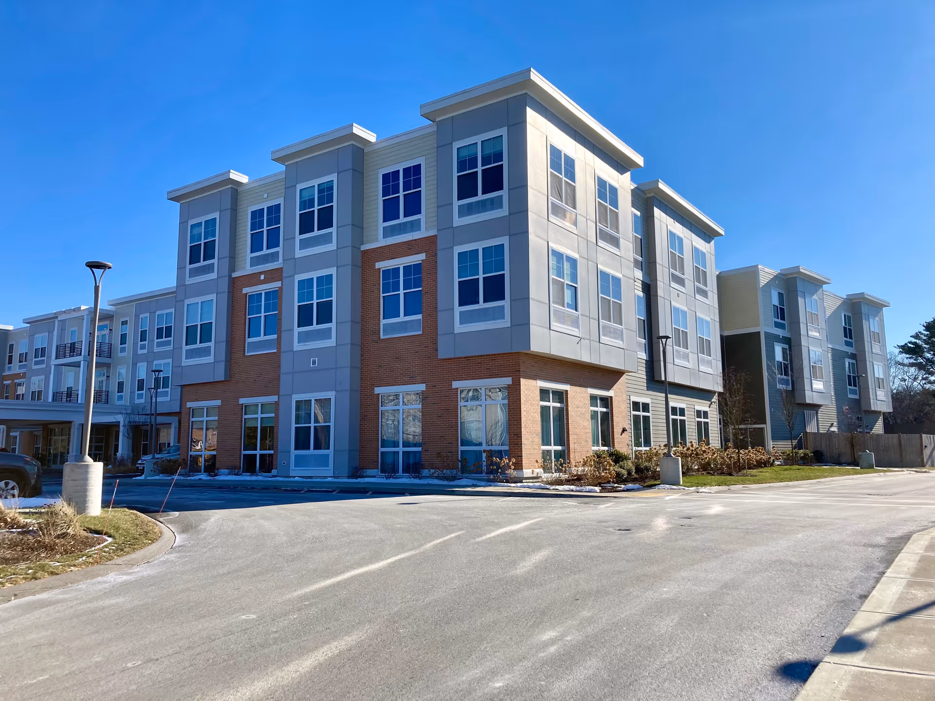 Front exterior of a three-story modern senior living building with brick and gray paneling and a driveway under a clear blue sky.