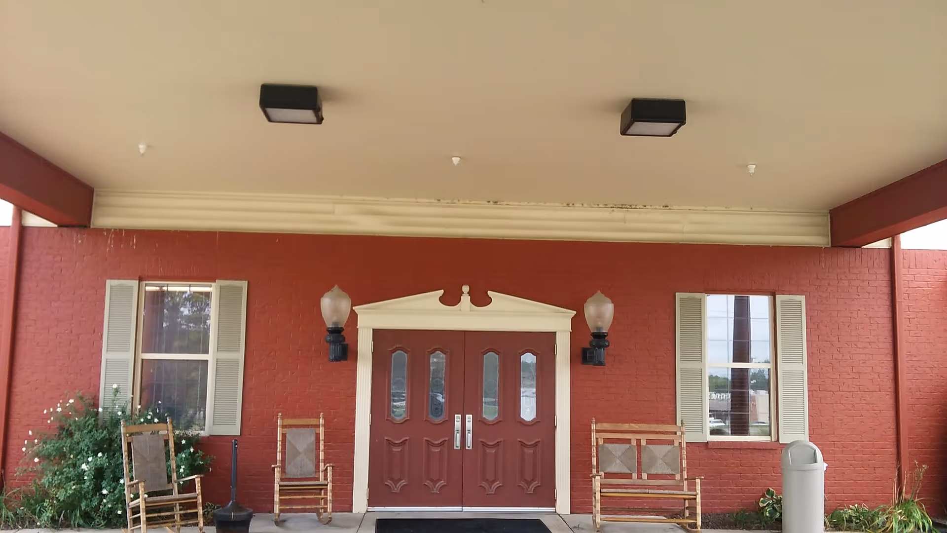 Red brick facility exterior with double doors, windows, benches, and trash can under a covered entrance.