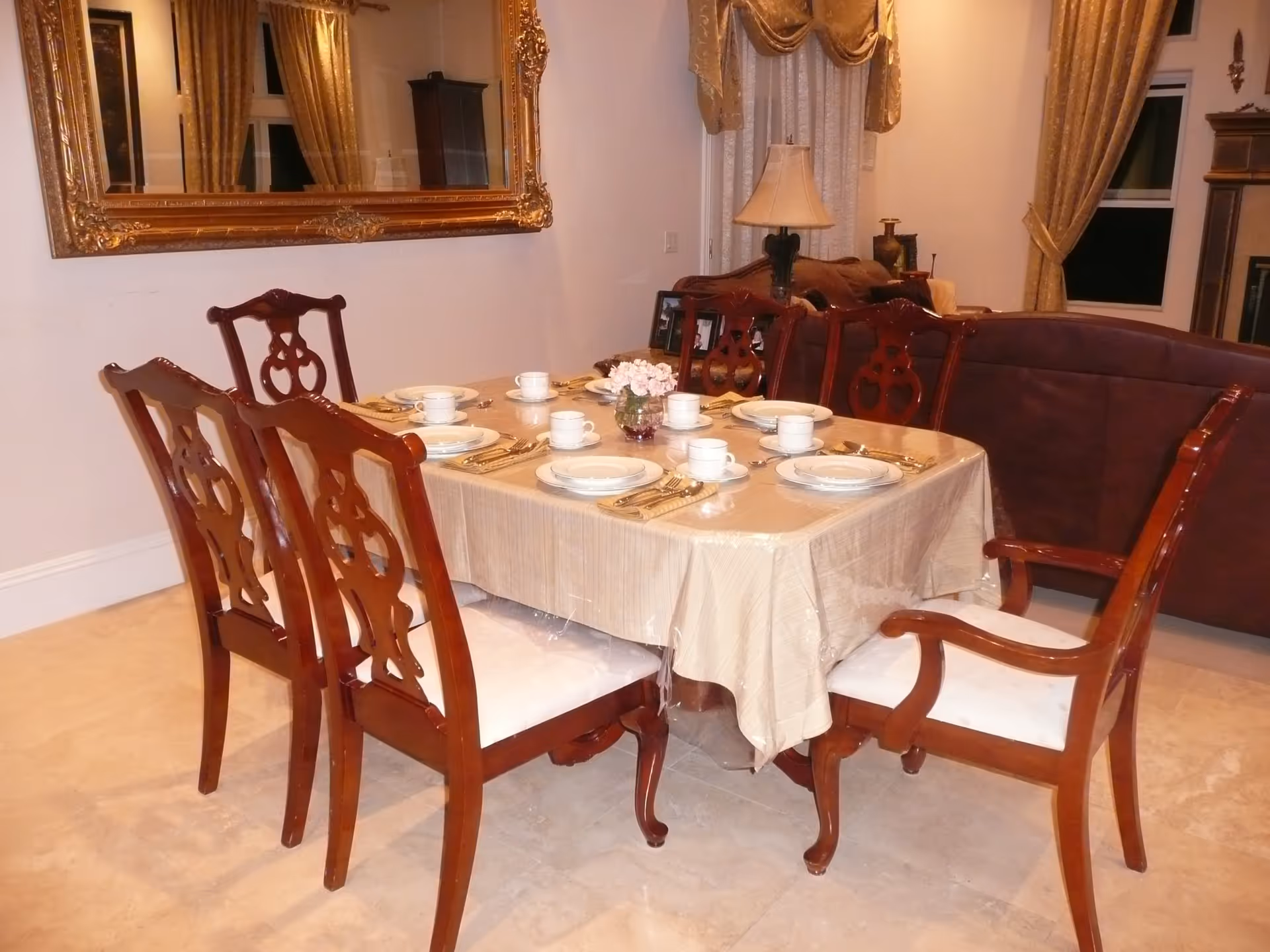 A dining room with a rectangular table covered with a beige tablecloth and set for six people with white plates, cups, and silverware. The table is surrounded by six wooden chairs with white cushions. Behind the table is a brown sofa, a lamp on a side table, and a large ornate gold-framed mirror on the wall. The room has beige curtains and a tiled floor.