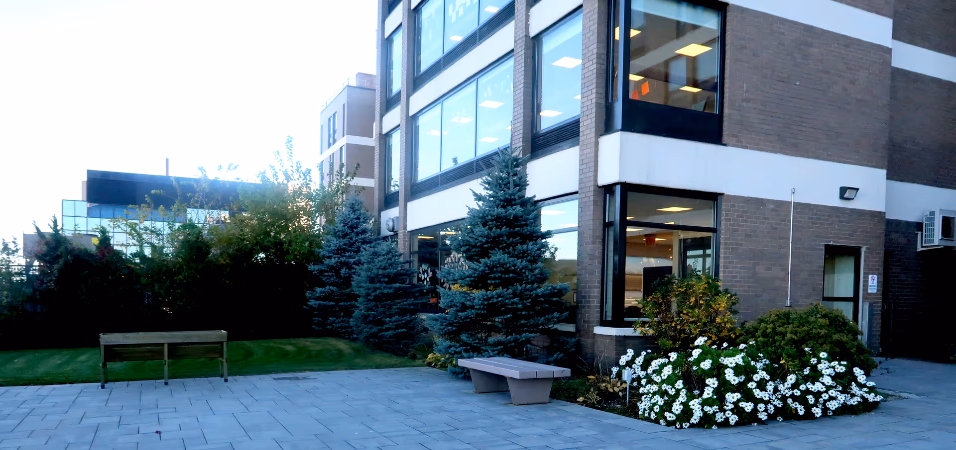 Exterior view of a brick facility with large windows, trees, benches, and flower beds.