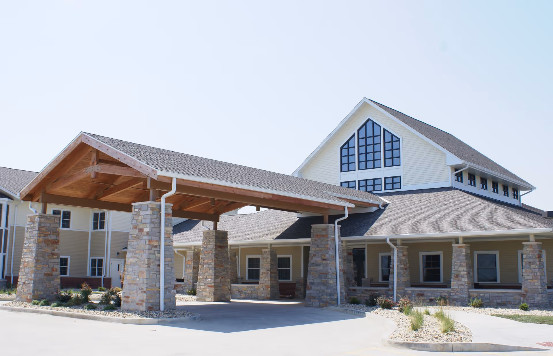 Exterior view of a senior living facility building with a covered entrance supported by stone pillars and a large windowed gable above the entrance area.