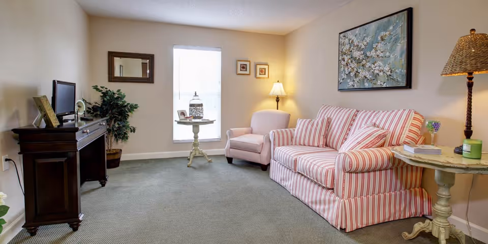 Bright furnished living room with a striped sofa and armchair, side tables, a TV console, and wall art.