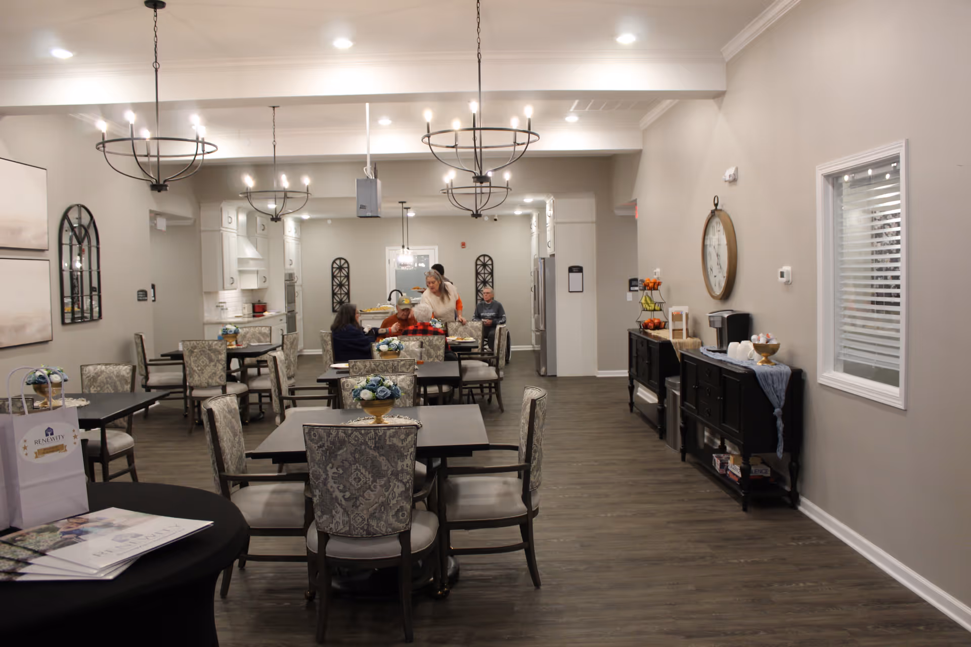A bright dining room with multiple tables, chairs, and staff serving residents.