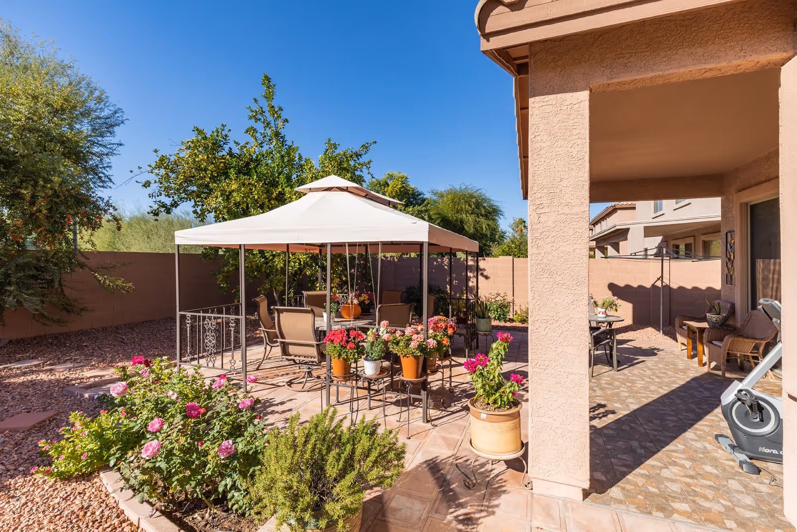 Outdoor patio area with a white canopy covering a table and chairs, surrounded by potted flowers and plants. The patio is adjacent to a beige stucco building with additional seating and an exercise bike visible under a covered area. The space is enclosed by a brick wall and features a clear blue sky.