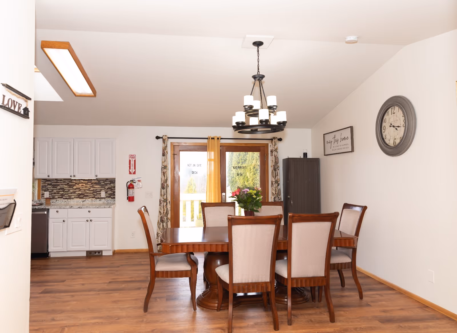 Dining room with wooden table and chairs, chandelier, clock, and adjacent kitchen area.