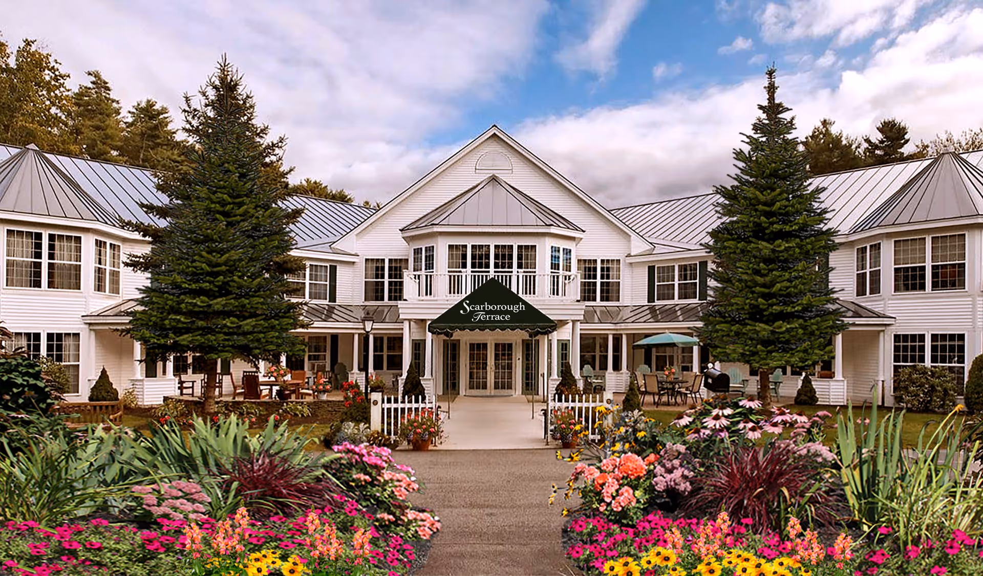 Front exterior view of Scarborough Terrace, a two-story white building with a metal roof, surrounded by tall evergreen trees and colorful flower beds along the walkway leading to the entrance.