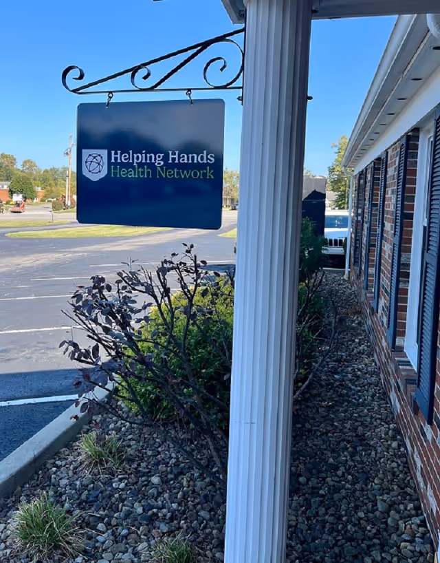 Blue hanging sign for Helping Hands Health Network beside a brick building exterior.