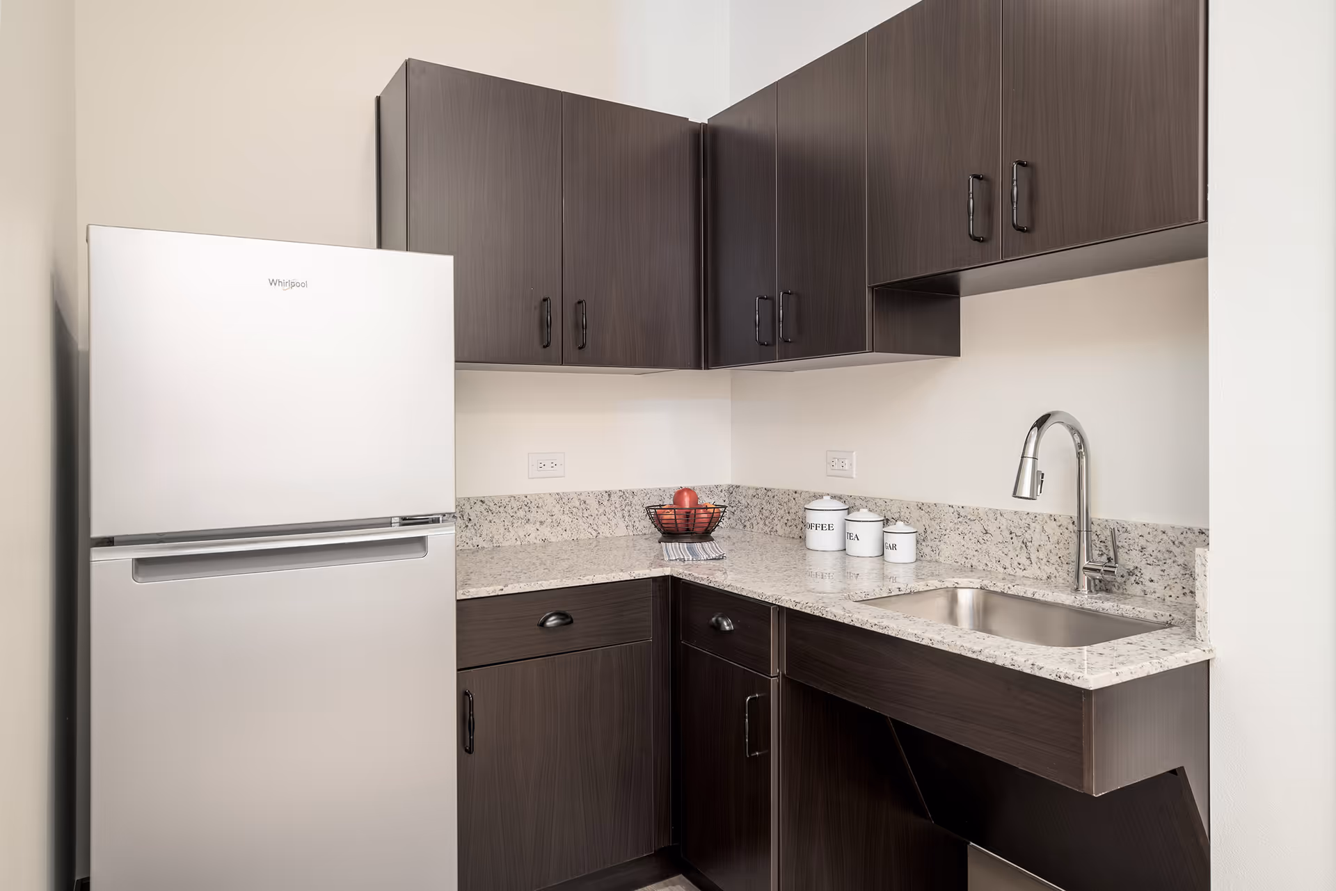 Compact kitchen area with dark wood cabinets, granite countertops, and a stainless refrigerator.