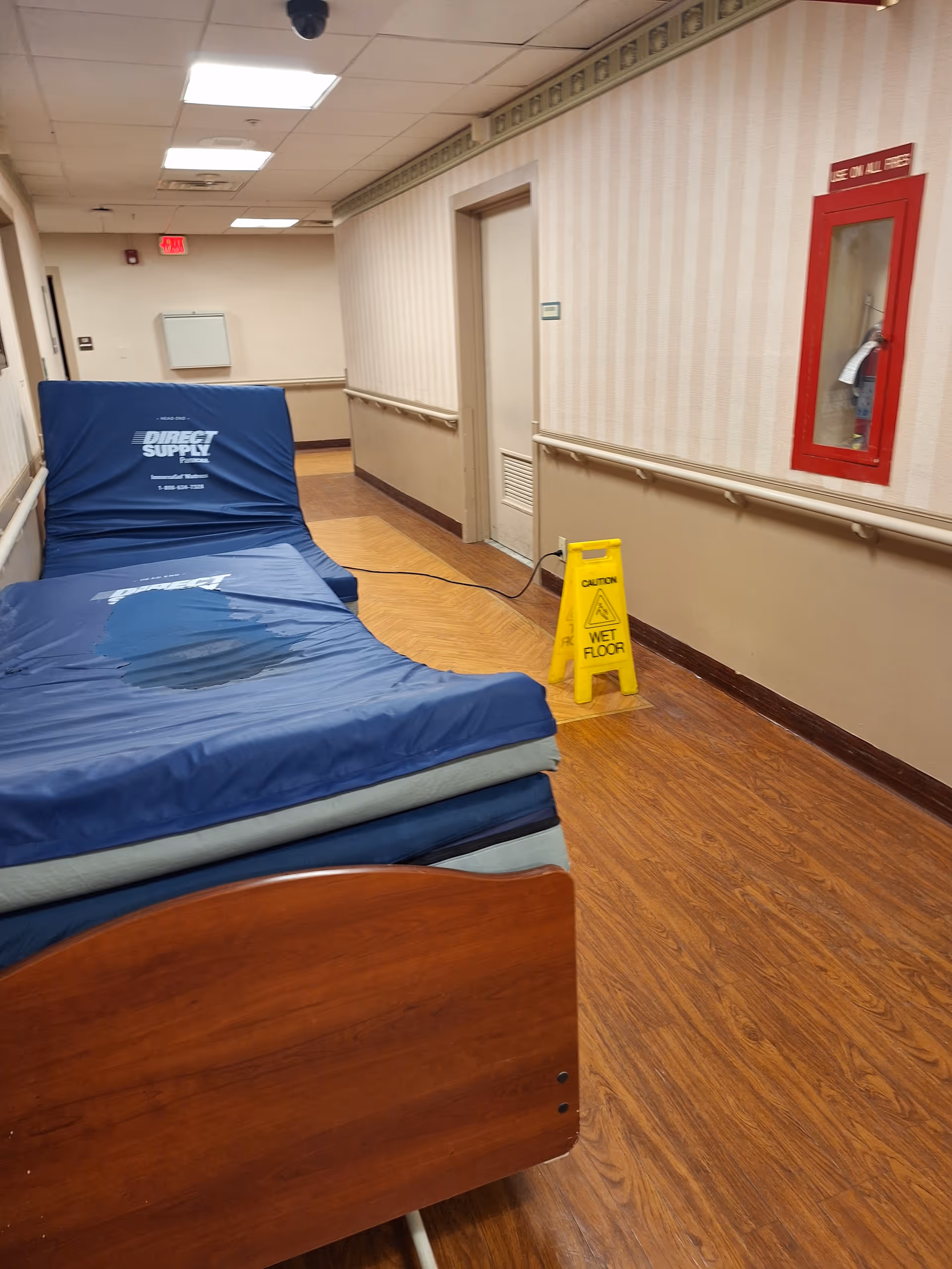 Narrow interior corridor with stacked blue mattresses, a wet floor sign, and a red wall cabinet.