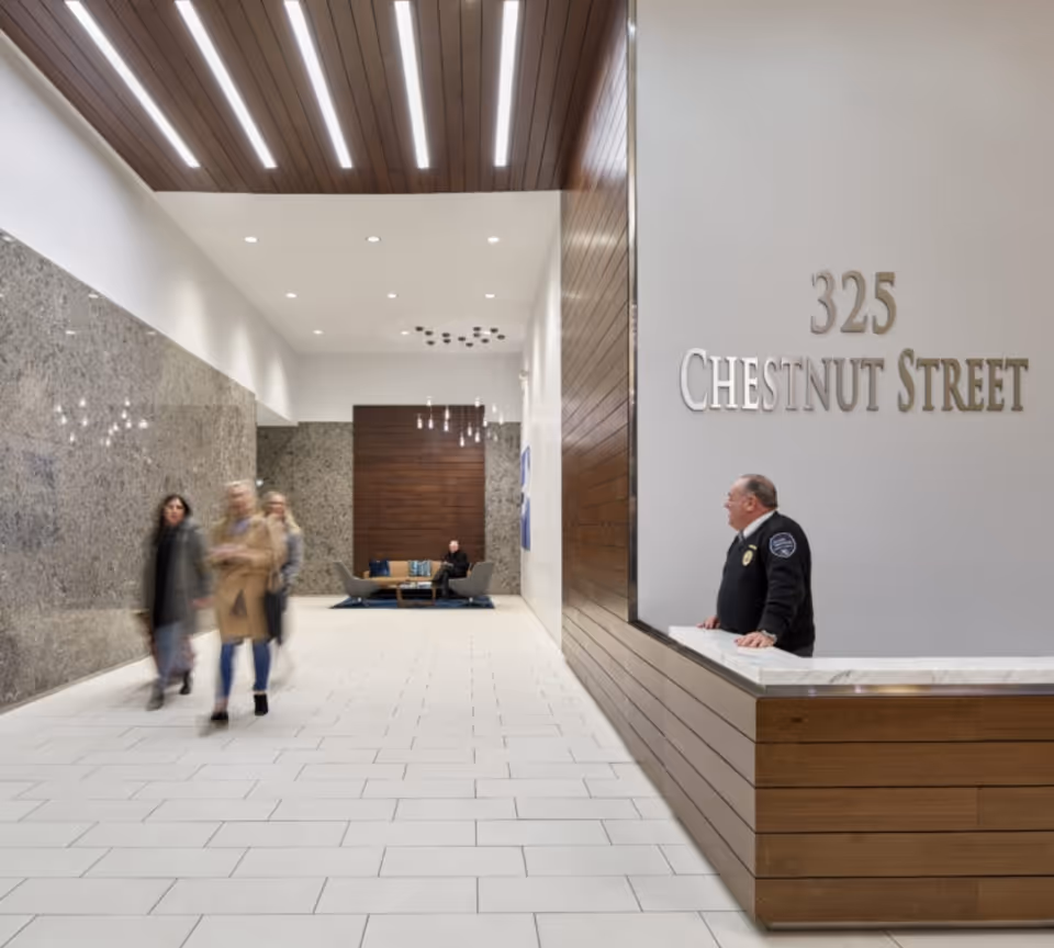 Modern interior lobby with a reception desk, wood-paneled ceiling, and blurred pedestrians passing.