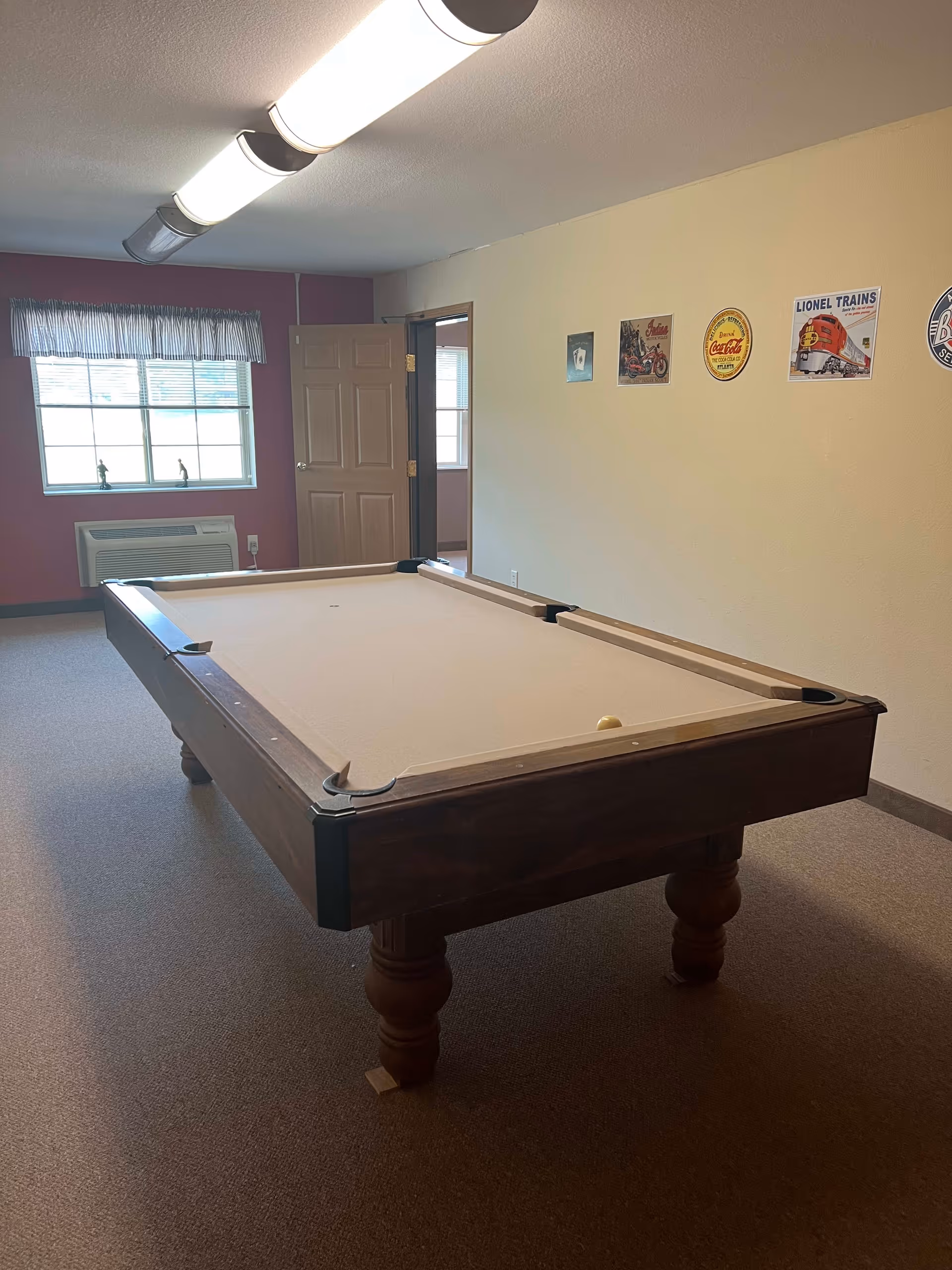 A room with a pool table in the center, beige carpet, and a beige wall with vintage-style signs including Coca-Cola and Lionel Trains. There is a window with a valance and an air conditioning unit below it. A door is open leading to another room.