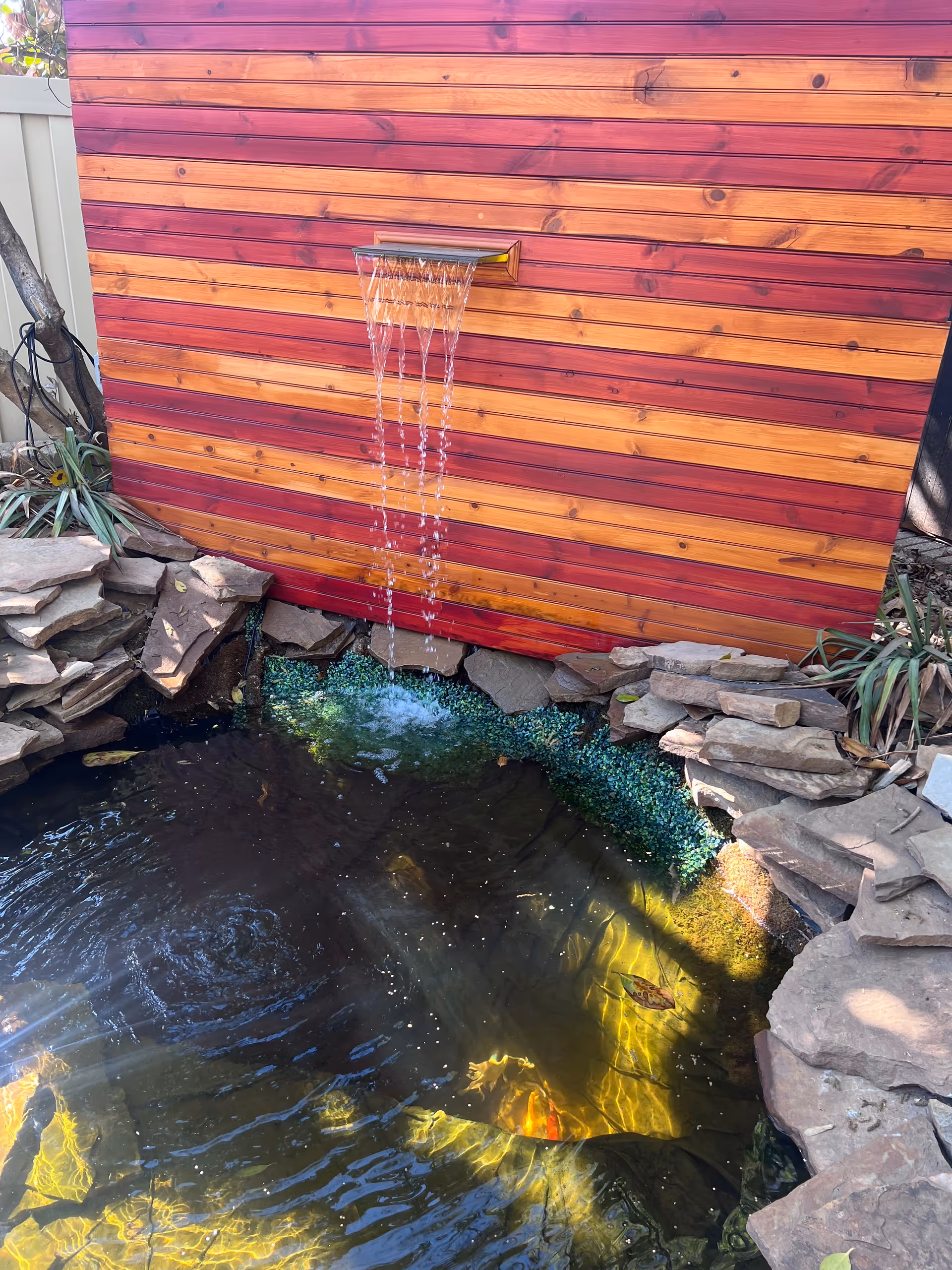 Wooden red-brown exterior wall with a metal water fountain pouring into a stone-bordered pond.