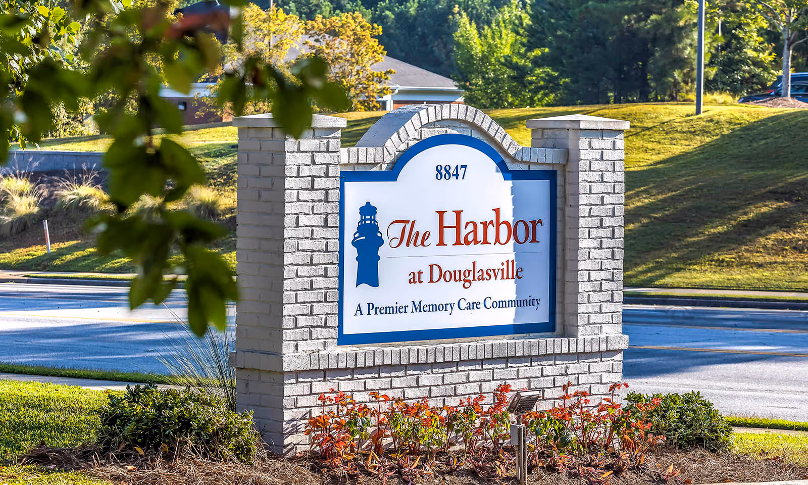 A brick sign for The Harbor at Douglasville, a premier memory care community, situated outdoors with greenery and a road in the background.