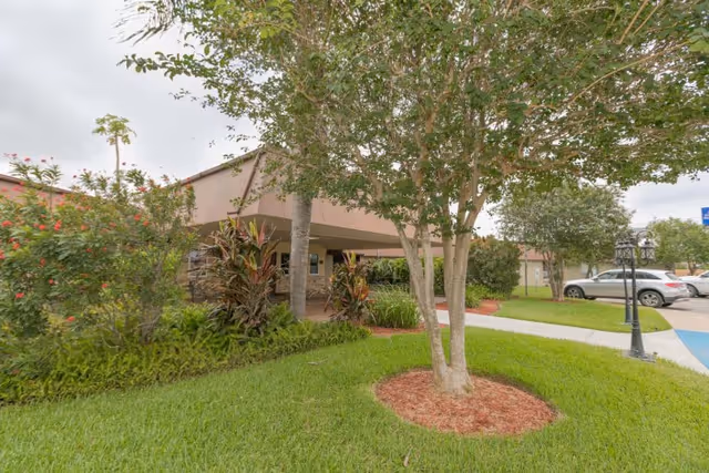 Landscaped front entrance of a single-story rehabilitation and healthcare building with trees, shrubs, a walkway, and parked cars.