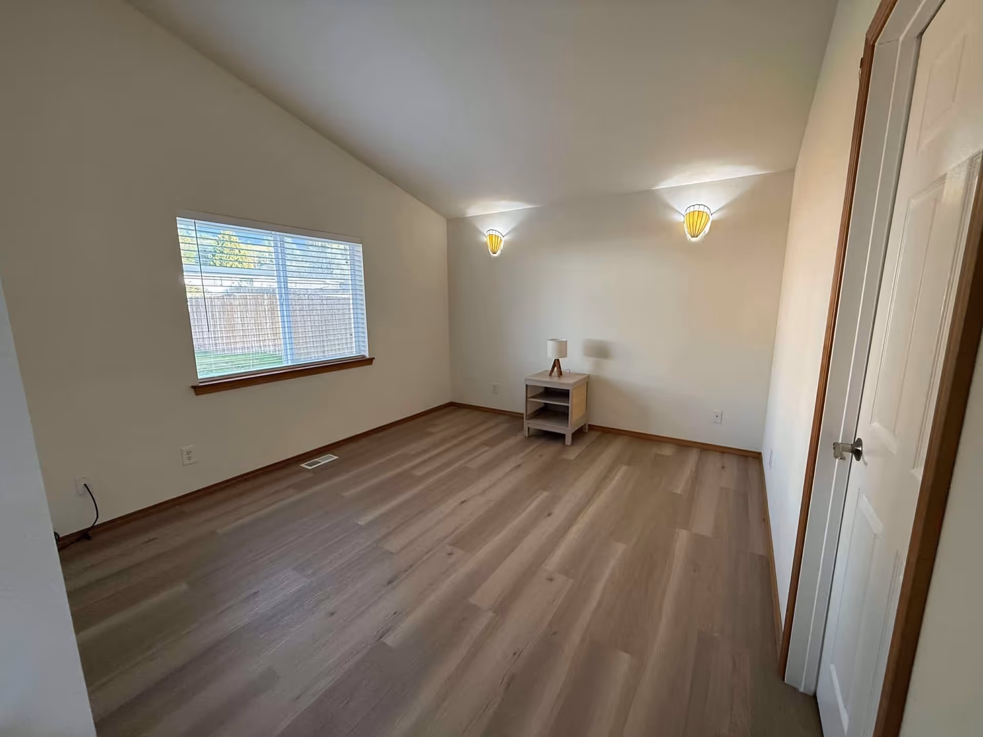 An empty room with wood floors, a window, and a small side table with lamp.