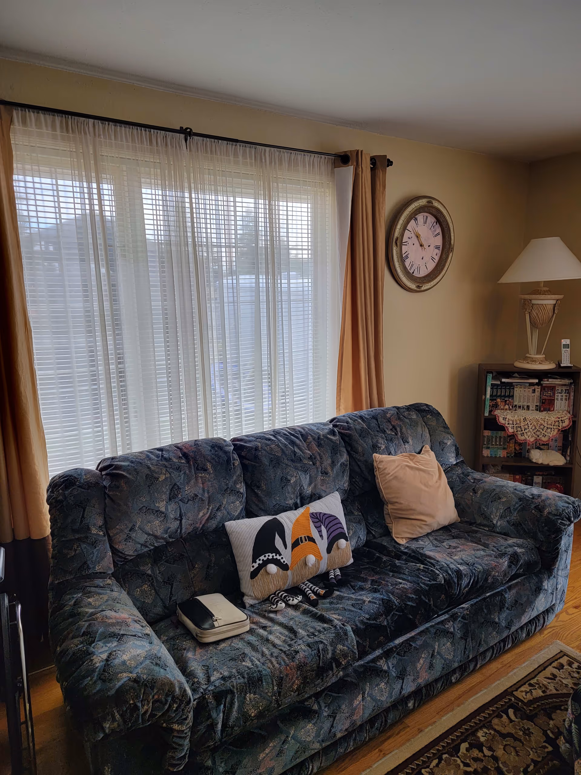 A cozy living room with a patterned blue couch featuring two pillows, one beige and one with a design of three gnomes. Behind the couch is a large window with sheer white curtains and beige drapes. On the wall to the right is a round clock and a small bookshelf with books and a lamp on top.
