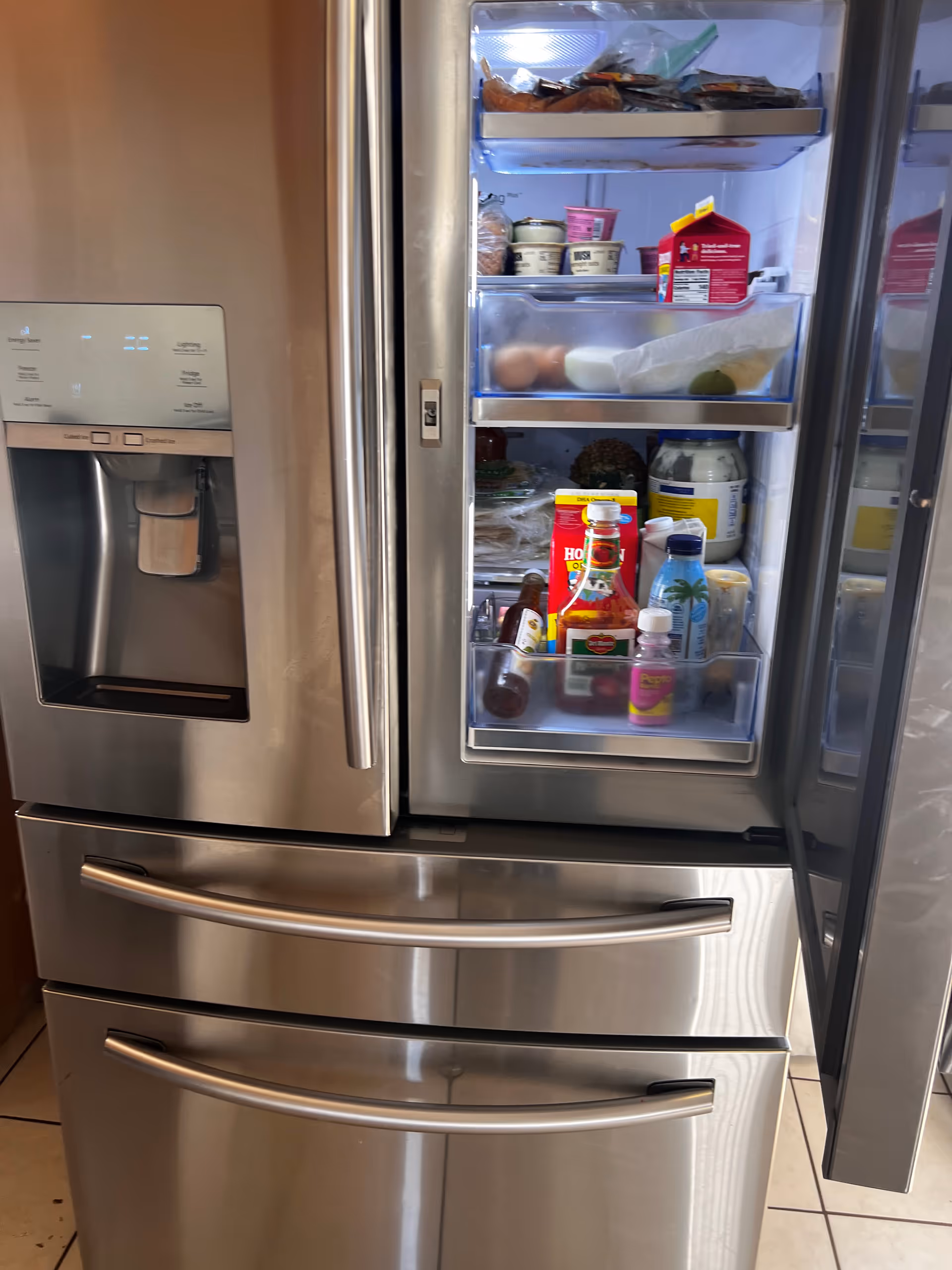 Open stainless steel refrigerator with ice dispenser and stocked shelves of groceries
