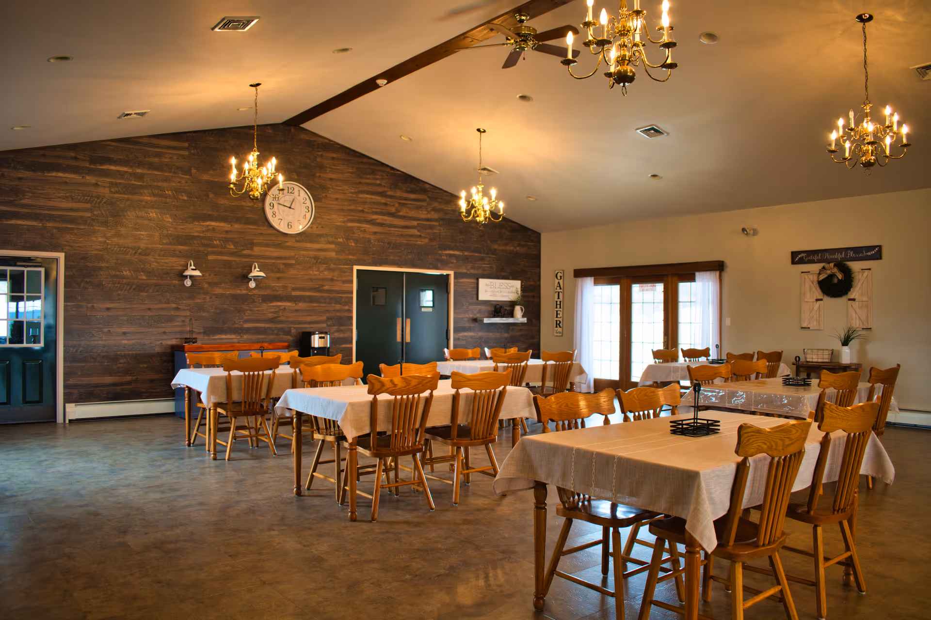 Spacious dining hall at Renee's Personal Care Home with wooden tables, chairs, chandeliers, and a clock.