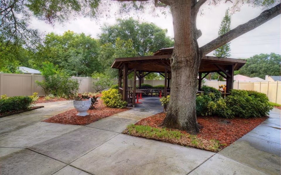 Outdoor area with a large tree in the foreground, surrounded by landscaped mulch beds and bushes. A wooden gazebo with benches and tables is situated in the middle, with a concrete pathway leading to and around it. The area is enclosed by a light-colored fence, and there are trees and greenery in the background.