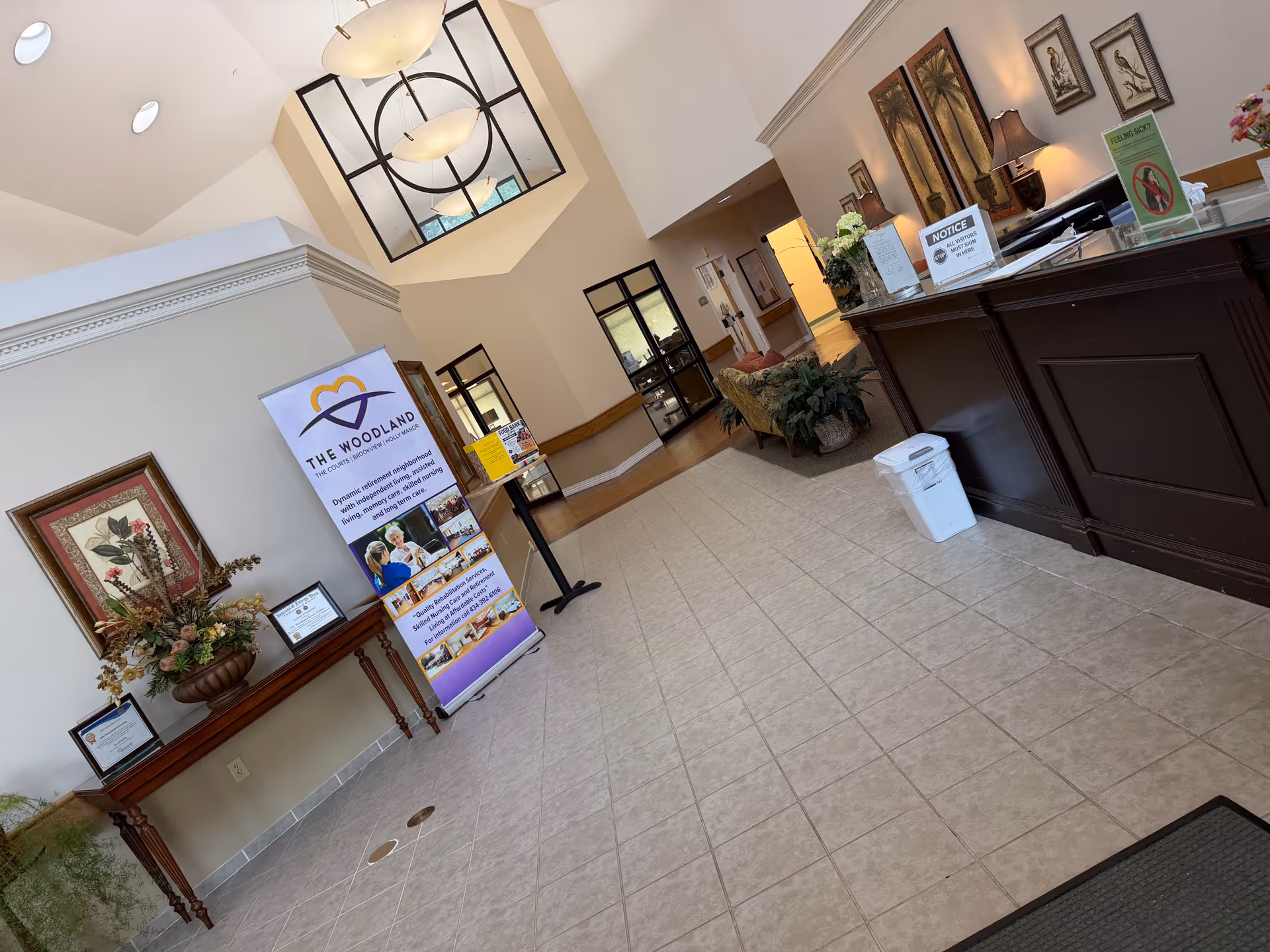 Senior living facility lobby with reception desk, banner stand, plants, and tiled floor.