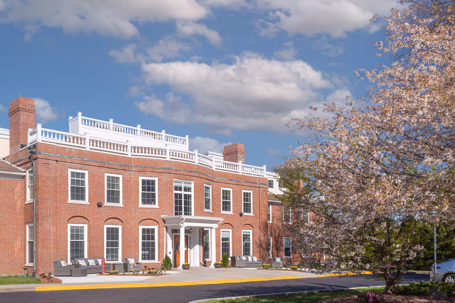 Brick senior living facility exterior with white railings, blooming tree, and outdoor seating.