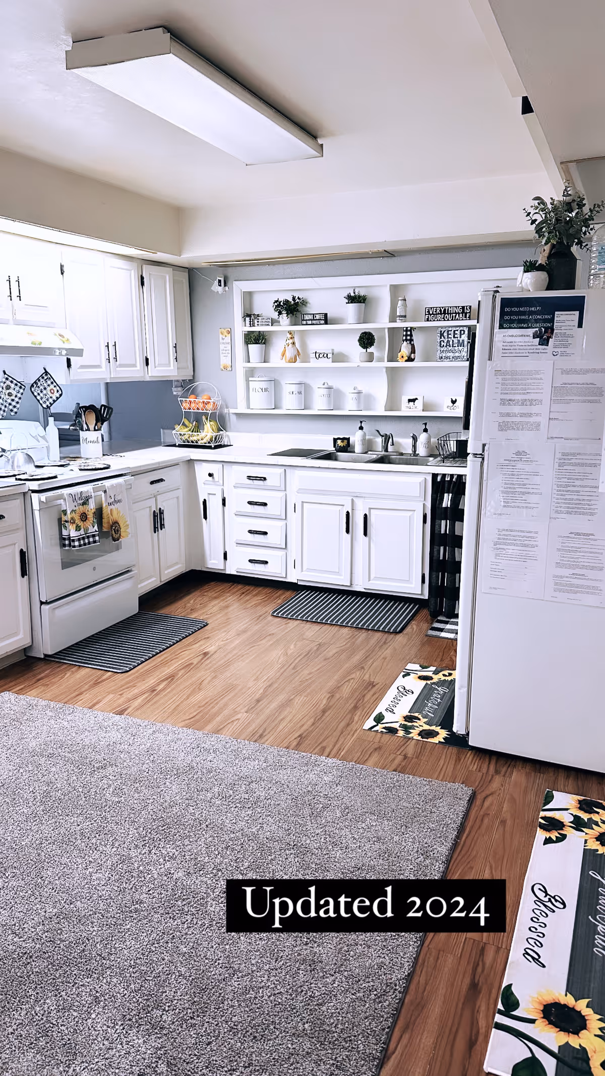 A bright and clean kitchen with white cabinets and drawers, a white stove with sunflower-themed towels, a white refrigerator covered with papers, and open shelves displaying decorative items and containers. The floor is a combination of wood and carpet, with striped and sunflower-themed mats placed around the kitchen area.