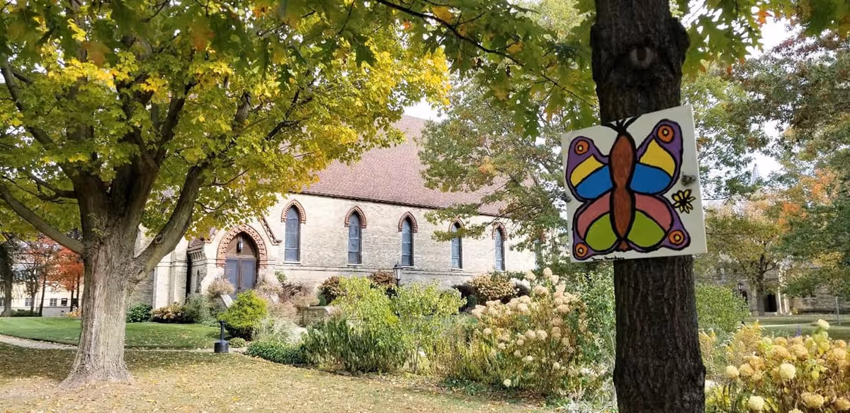 Stone building with arched windows and a red roof viewed across a lawn and trees, with a colorful butterfly drawing attached to a foreground tree.