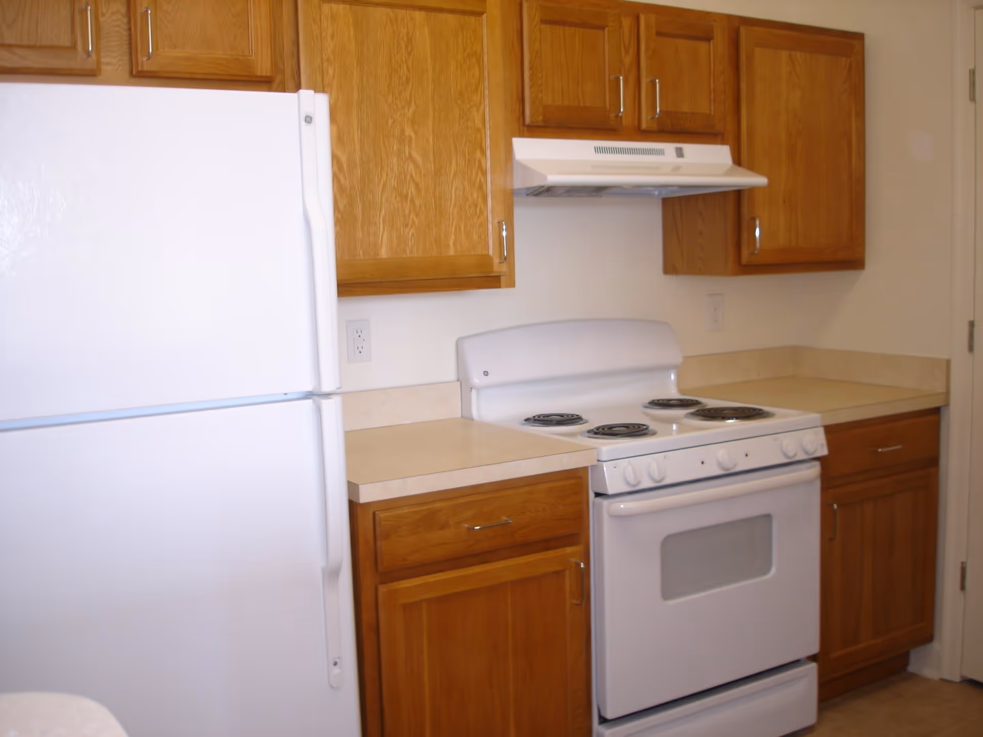 Small kitchen with a white refrigerator, white electric stove, wooden cabinets and beige countertops.