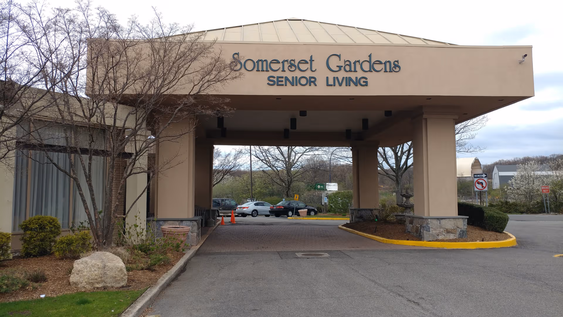 Exterior view of Somerset Gardens Senior Living entrance with driveway, sign, and parked cars.