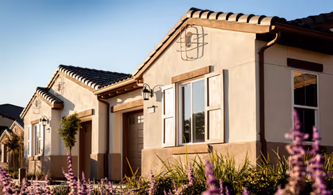 Exterior view of a single-story residential building with beige stucco walls, brown tiled roof, and wooden shutters on the windows. There are plants and purple flowers in the foreground under a clear sky.