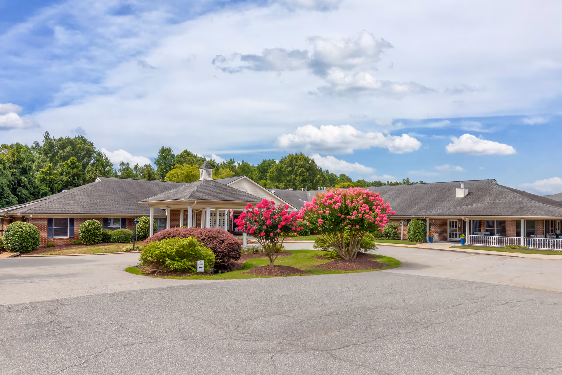 Single-story brick senior living facility with central entrance, circular driveway, and flowering shrubs.