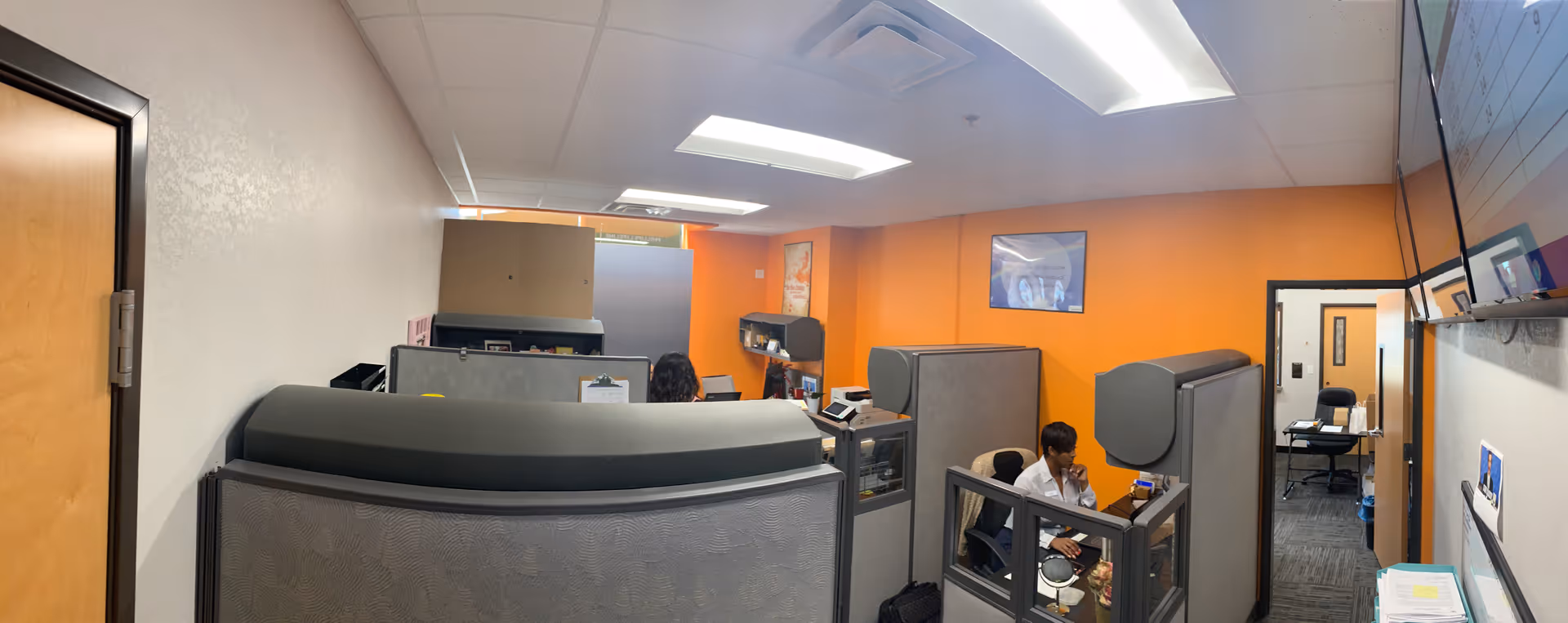 Office cubicles with staff working at desks in an orange-walled administrative area.