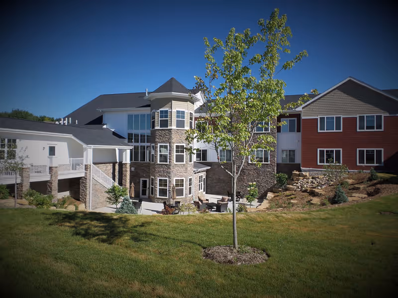 Front exterior of a multi-story senior living building with stone and siding facades, patio seating and a landscaped lawn with a young tree.